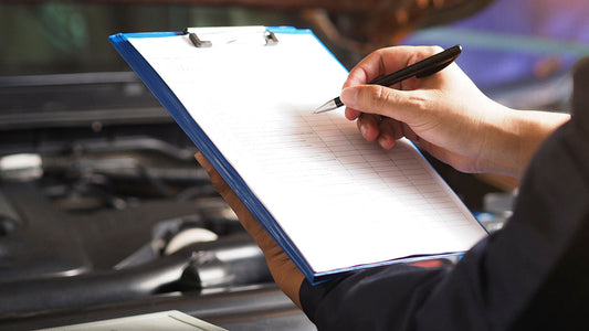 A printed collision repair estimate on a clipboard showing part line items with abbreviations, a pen resting beside it on a desk in an auto repair shop office