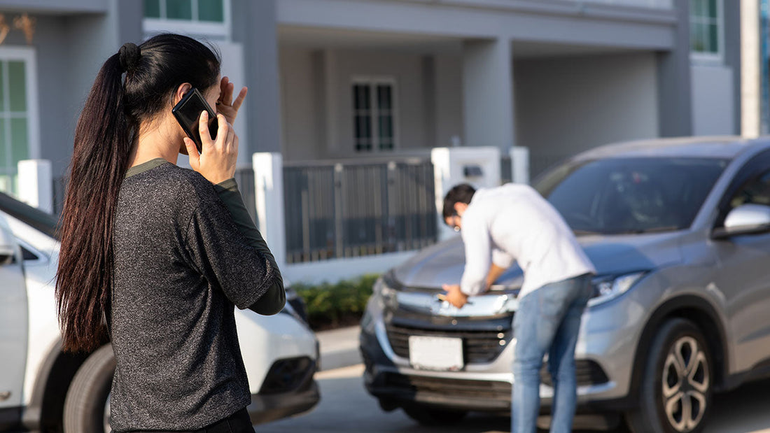 A driver standing safely on the shoulder beside a lightly damaged car, hazard lights on, making a phone call after a minor collision